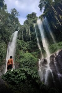 Explorer standing before a vibrant waterfall in Bali's lush rainforest.