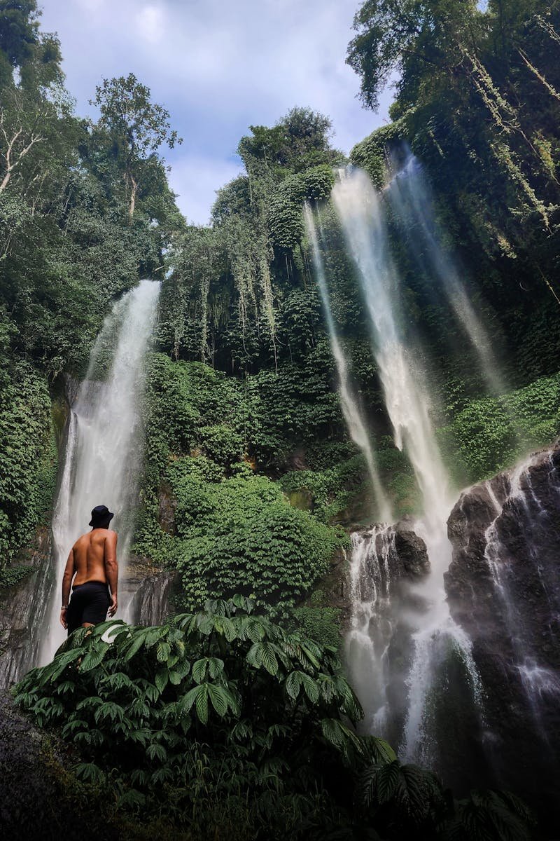 Explorer standing before a vibrant waterfall in Bali's lush rainforest.